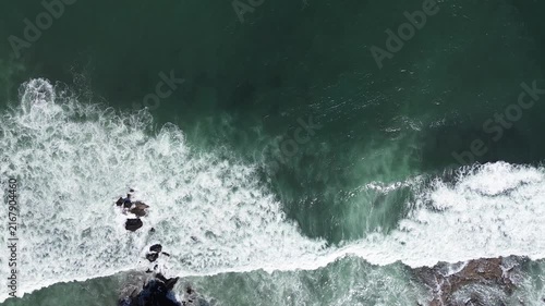 Aerial view shows waves crashing against rocks. Deep green water churns with white foam. Rocks jut from the sea, battered by surf. Nature's power is raw and relentless