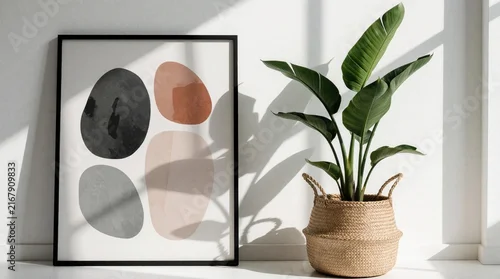 Panel kuchenny z motywem Abstract organic shapes art in black frame on white wall. Minimalist interior decor features potted plant in wicker basket. Natural sunlight creates soft shadows in modern living room. Stylish earth