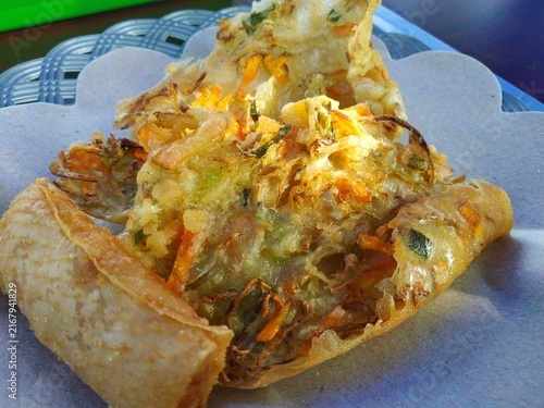 Close-up view of popular Indonesian traditional street food, featuring crispy vegetable fritter (bakwan) and fried spring roll (lumpia), served on brown food wrap paper over a woven plate.