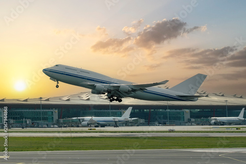 View of a commercial airline passenger airplane taking off from the runway, with the airport terminal building structures, against the backdrop of the rising sun and clouds