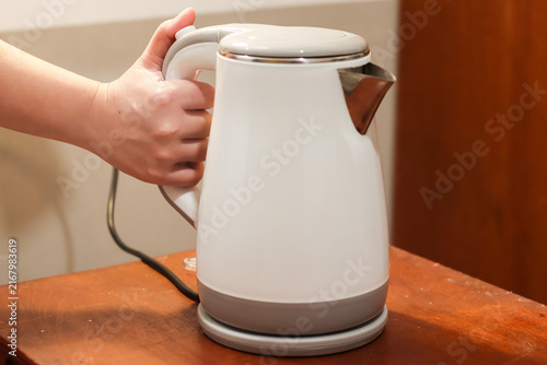Electric kettle on a wooden table, held by a woman's hand, used for boiling water in a kitchen, office pantry, or hotel room.