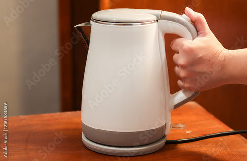 Electric kettle on a wooden table, held by a woman's hand, used for boiling water in a kitchen, office pantry, or hotel room.