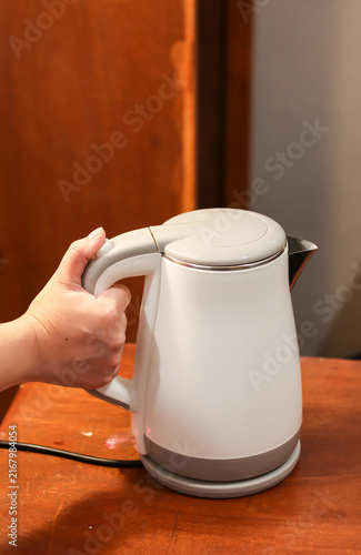 Electric kettle on a wooden table, held by a woman's hand, used for boiling water in a kitchen, office pantry, or hotel room.