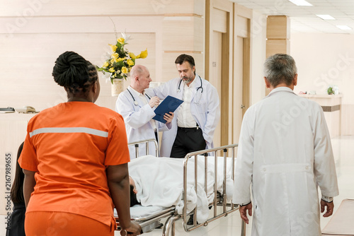 Doctors and a nurse in a hospital corridor wheel a patient on a stretcher while discussing notes, coordinating urgent care.