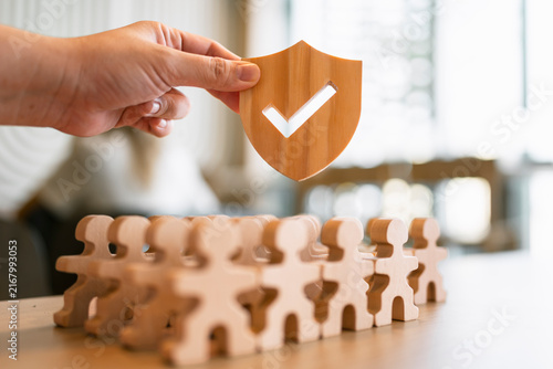 Fotografie Life insurance and employee protection concept with hand holding wooden shield with checkmark over people figures for healthcare coverage, social security guarantee and risk management
