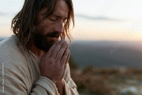 Man with long hair and beard clasps hands in quiet prayer. Soft golden light bathes his bowed head. Mountains stretch behind him under twilight sky. He rests on rugged terrain, deeply still