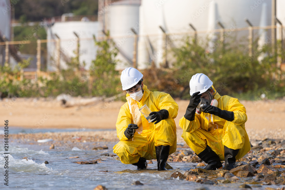 Fototapeta premium Serious environmental worker in hazmat suit inspecting polluted water at river edge alongside safety inspector with focused expression during daytime hazardous material spill testing