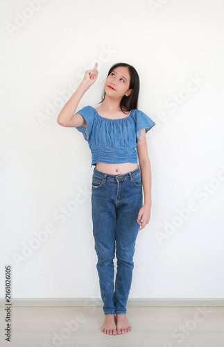 An Asian preteen girl wearing a blue denim crop top and jeans stands against a white background, looking up and pointing her index finger upwards.