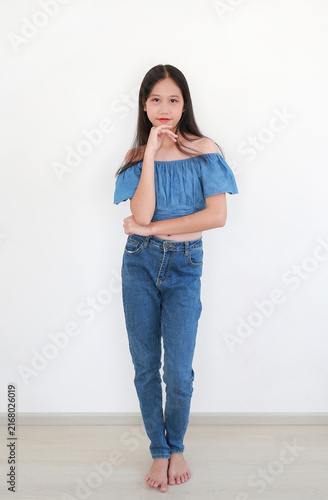 A young Asian girl wearing a blue off-shoulder crop top and jeans poses gracefully with her hand on her chin against a white wall.