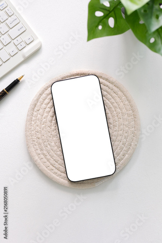 Top view flat lay of modern smartphone mockup with blank white screen on woven coaster, white desk, keyboard, pen, and green plant. Minimalist workspace template for aesthetic design.