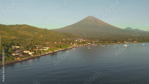 Aerial view of the tropical coastline of Bali with Mount Agung volcano rising in the background, showing a coastal village, ocean, and lush green landscape.