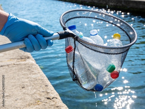 A person in blue gloves lifts a net full of discarded plastic bottles from a shimmering canal.