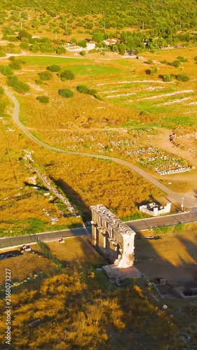 Vertical video. Gelemis, Turkey. Monumental triple-arched Roman triumphal gateway of Mettius Modestus, weathered stone structure spanning a modern road amid golden fields and distant hills, aerial vi