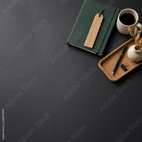 Top view of a dark desk with a green notebook, coffee cup, and stationery accessories
