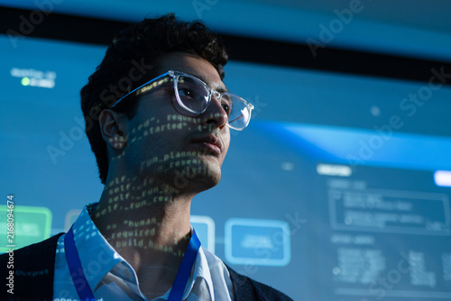 Technology Specialist Focused on System Monitoring with Digital Dashboard Reflected Across Face. software engineer focused on system analytics with digital data standing in control room at tech office