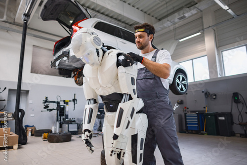A man in overalls works on a humanoid robot in a car repair shop with a vehicle lifted overhead