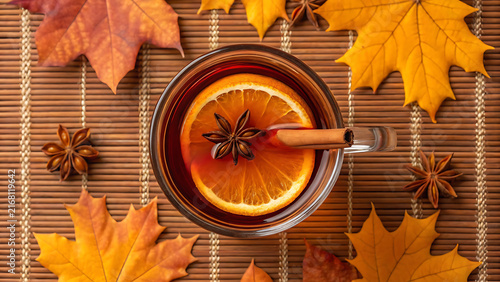 Autumn tea in glass cup with cinnamon stick and orange slice on wooden table with fall leaves