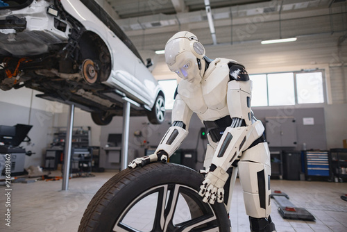 A humanoid robot with glowing blue eyes is carefully handling a car tire in a repair shop setting