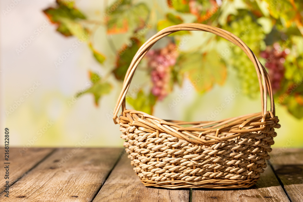Fototapeta premium Empty wicker basket on rustic wooden table with grape leaves behind for autumn harvest product scene