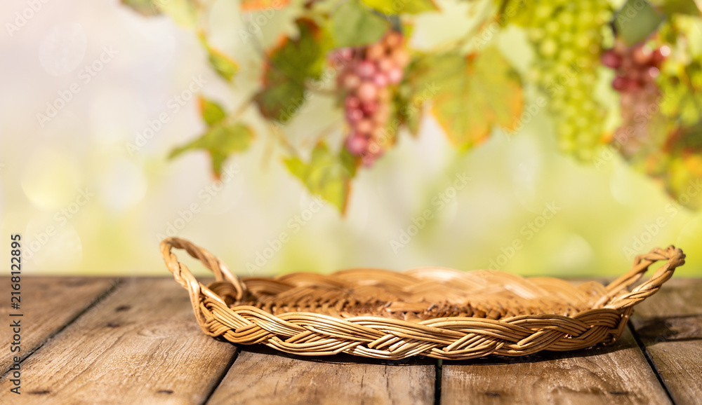 Fototapeta premium Flat wicker tray on wooden table with blurred grapes and vine leaves for rustic autumn product background