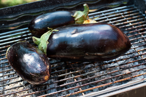 Fresh whole eggplants are grilling on a hot metal barbecue grate outdoors