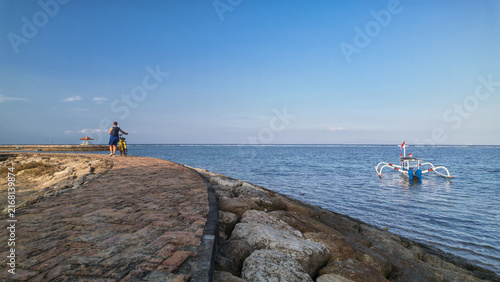 Foto Person Walking with Bicycle on Sanur Beach Stone Breakwater Jetty Near Tradition