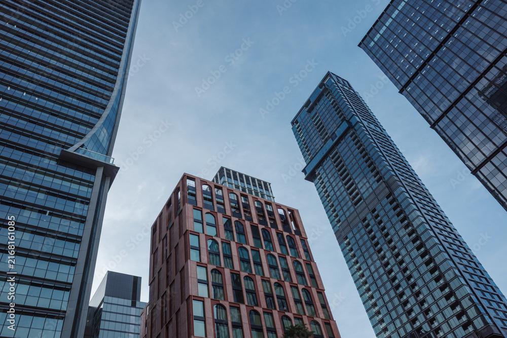 Fototapeta premium Low-angle view of multiple modern corporate skyscrapers featuring glass and brick architectural facades under a clear blue sky during the day.