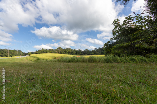 A low-angle landscape view of a green grassy field leading to rolling hills and trees under a vibrant cloudy sky.
