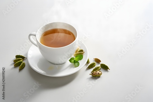 A steaming white ceramic cup of aromatic cardamom tea served on a saucer with fresh green mint leaves and whole cardamom pods scattered on a clean white background.