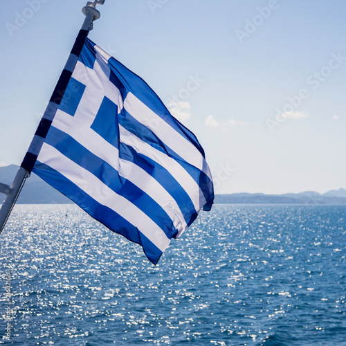 Greek flag billowing from ship above sparkling Aegean Sea water, blue sky, Greece