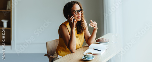 Woman sits at office table talking on the phone with coffee and notebook