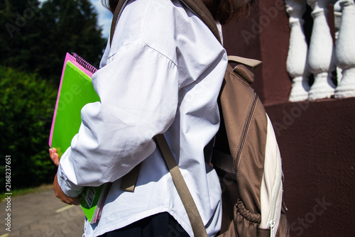Schoolgirl stands in front of a school holding notebooks and supplies while wearing a backpack. It is back to school season