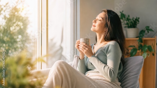 Calm young woman enjoying hot drink while relaxing by bright sunny window at home