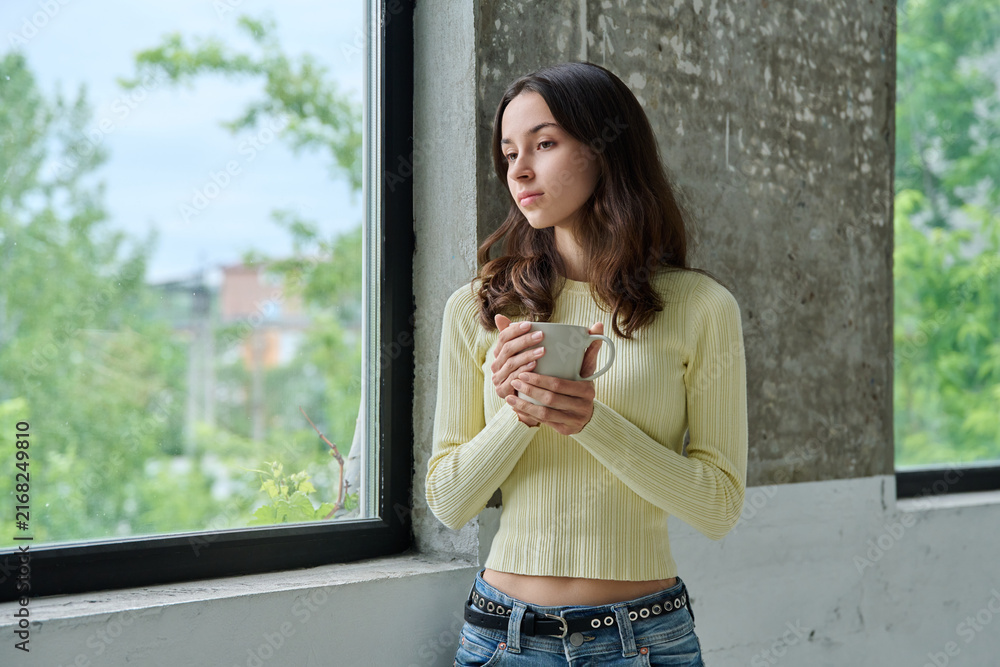 Fototapeta premium Pensive young woman holding mug standing near window and looking outside