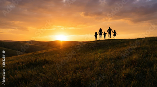 Silhouette of happy family of five running on grassy hill crest during golden sunset with dramatic clouds in wide rural landscape.