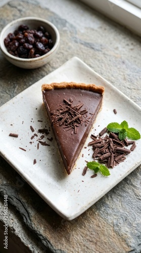 A slice of rich chocolate tart on a square plate, garnished with chocolate shavings and fresh mint leaves.