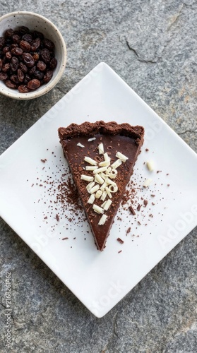 Slice of chocolate tart with white chocolate shavings on a plate, bowl of raisins on stone background.
