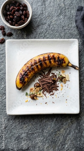 Grilled banana dessert with chocolate shavings and almonds, gourmet fruit platter on dark stone surface.