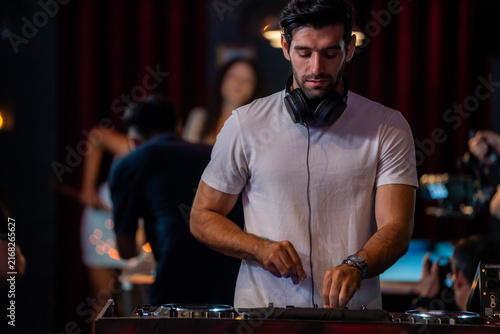 Obraz na płótnie A wide view of a male DJ standing behind turntables and a mixer in a busy nightclub reaches for the pitch fader in a wash of red light in a wide nightclub interior with pool tables
