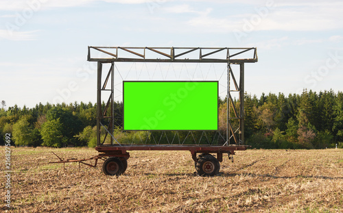 Large green chroma key billboard mounted on a metal trailer stands in a harvested rural field, with trees and open sky in the background