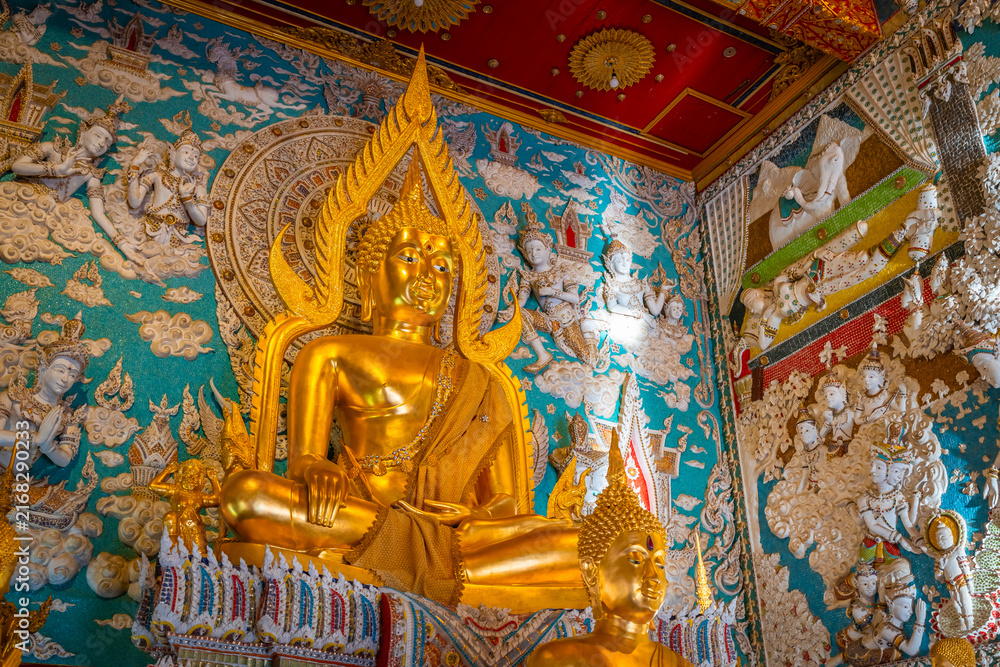 Fototapeta premium Golden Buddha statue inside the hall of Wat Sri Uthumphon in Mueang Nakhon Sawan District, Nakhon Sawan, Thailand, surrounded by ornate white relief wall art and celestial angels.