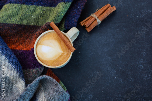 Cup of spicy coffee with milky foam, cinnamon sticks, warm scarf on black background. Top view with copy space. Autumn or winter mood, seasonal hot drink