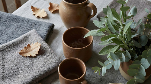Autumnal Gathering: Cozy ceramic mugs and textured linen napkins are artfully arranged on a rustic table, accented with dried oak leaves and fresh eucalyptus. A perfect setting for a warm.