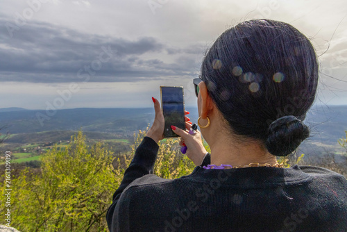 Cuadro en lienzo A woman from behind holding a smartphone to capture a scenic view of rolling green hills under a cloudy sky