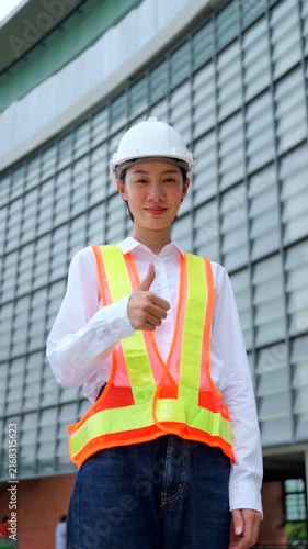 Smiling Asian woman civil engineer in safety vest showing thumbs up sign outside corporate office building. Professional contractor project success concept. Vertical Shot