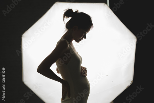 Картината върху платно Silhouette of a pregnant woman in profile, gently cradling her belly against a bright, octagonal light background