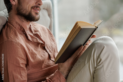 Happy young man smiling while reading a book by a window during leisure time