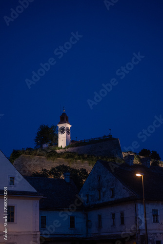 Illuminated historic clock tower stands atop a Petrovaradin fortress wall at night