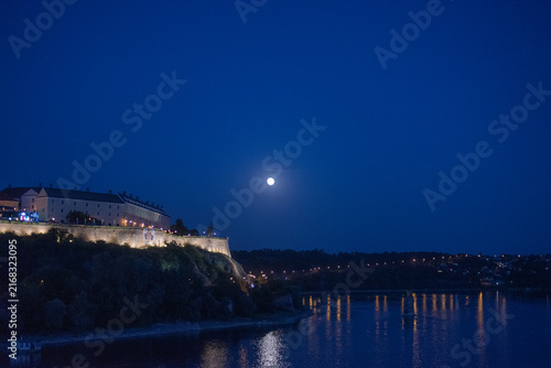Historic Petrovaradin fortress sits on a cliff overlooking a calm Danube river under a dark night sky with full moon