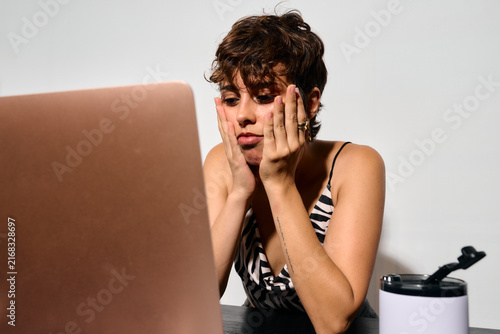 Bored student at home. DIrect flash shot. She stares intently at her laptop screen, hands pressed to her cheeks. Overwhelmed by stress, she looks exhausted and mentally drained.